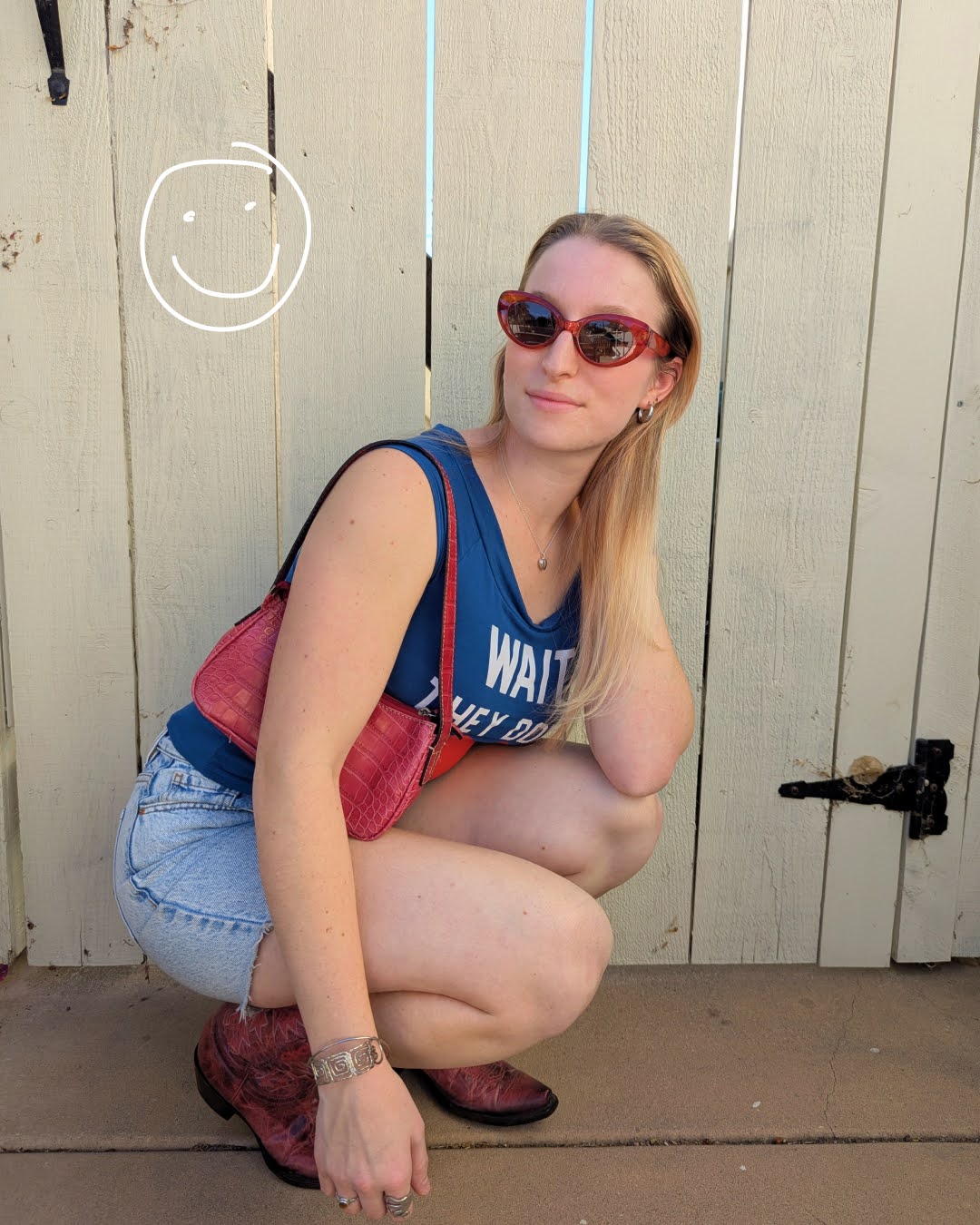 Grace, a white woman with blond hair, crouches down and smiles for a photo. She is wearing a navy blue top with light wash denim shorts, plus red cowboy boots, a red purse, and red sunglasses. A smiley face is drawn in white ink in the top left corner of the photo.